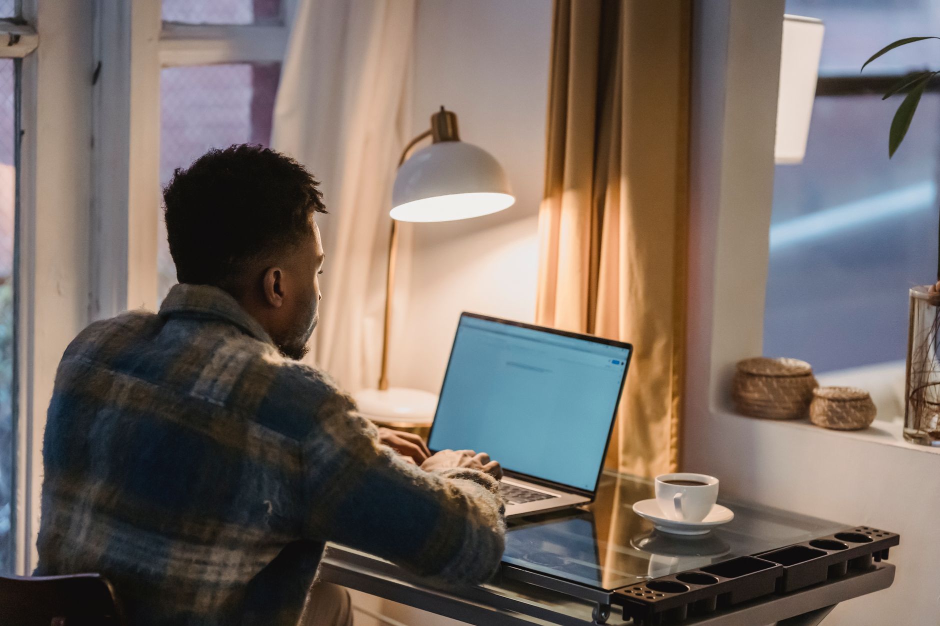 Man in een geruit overhemd zit 's avonds aan een bureau naast een lamp en typt op een laptop, met een kopje koffie binnen handbereik