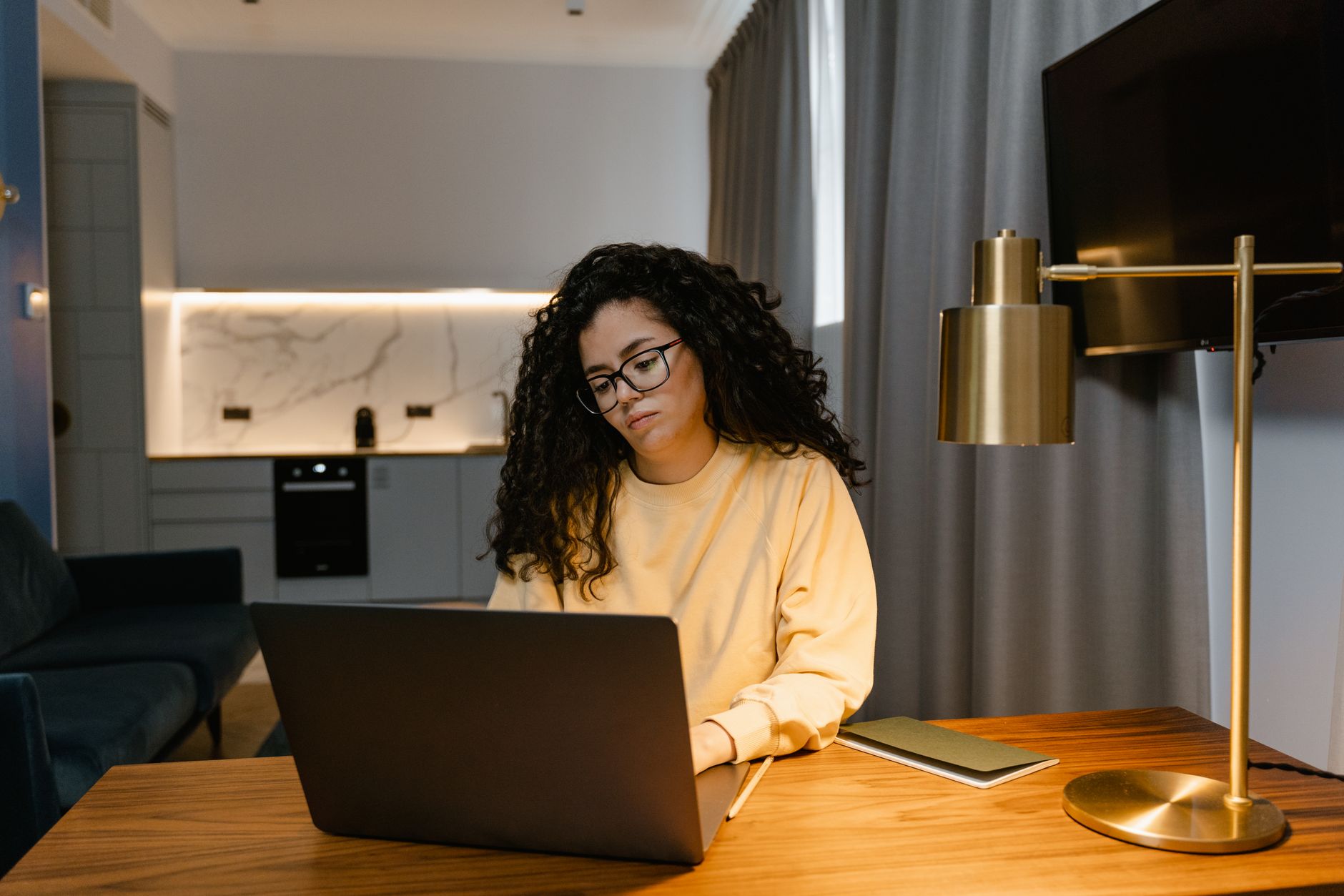 Vrouw met bril en gele trui werkt geconcentreerd op een laptop aan een houten tafel in een woonkamer