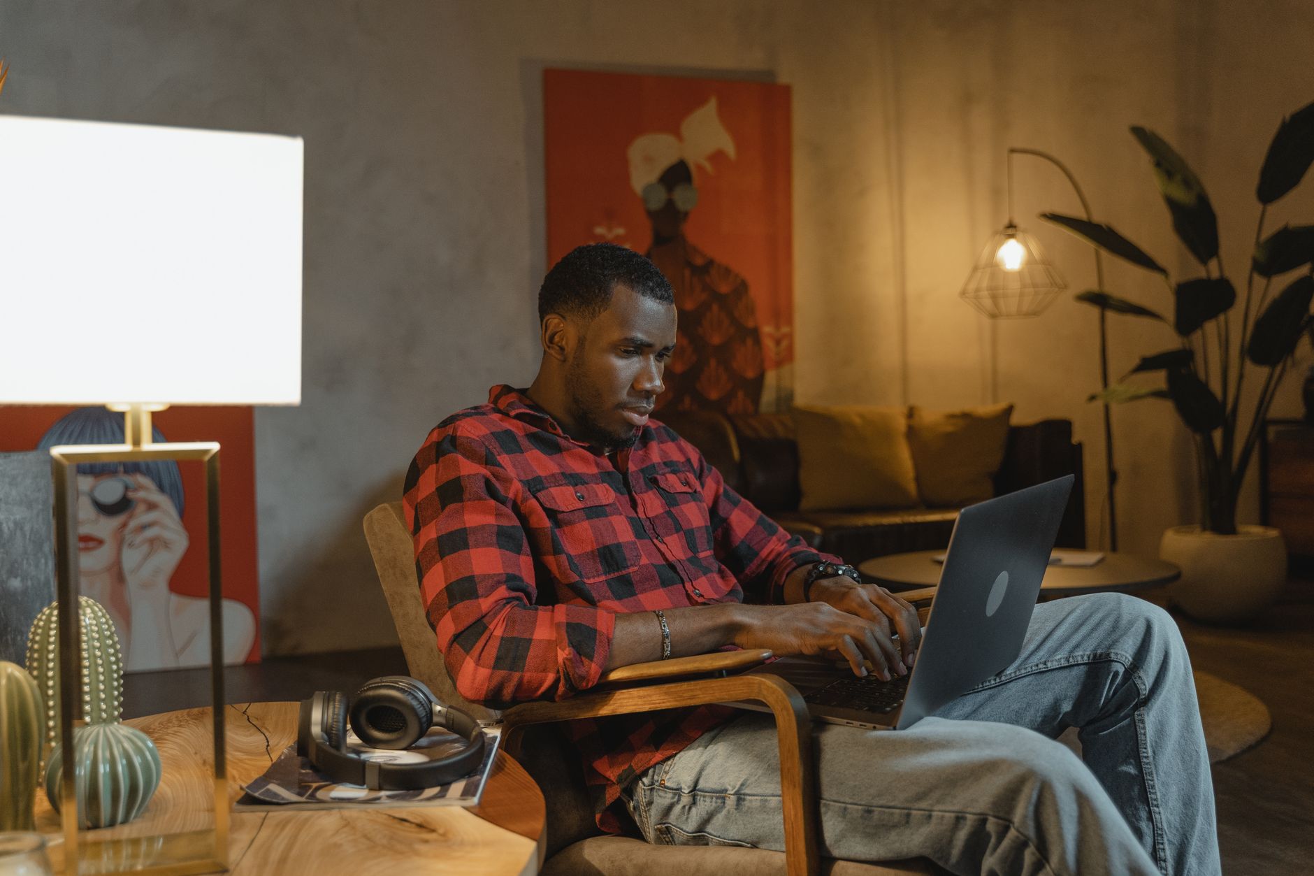 Man in een rood geruit overhemd zit in een fauteuil met een laptop op schoot in een gezellig verlichte woonkamer