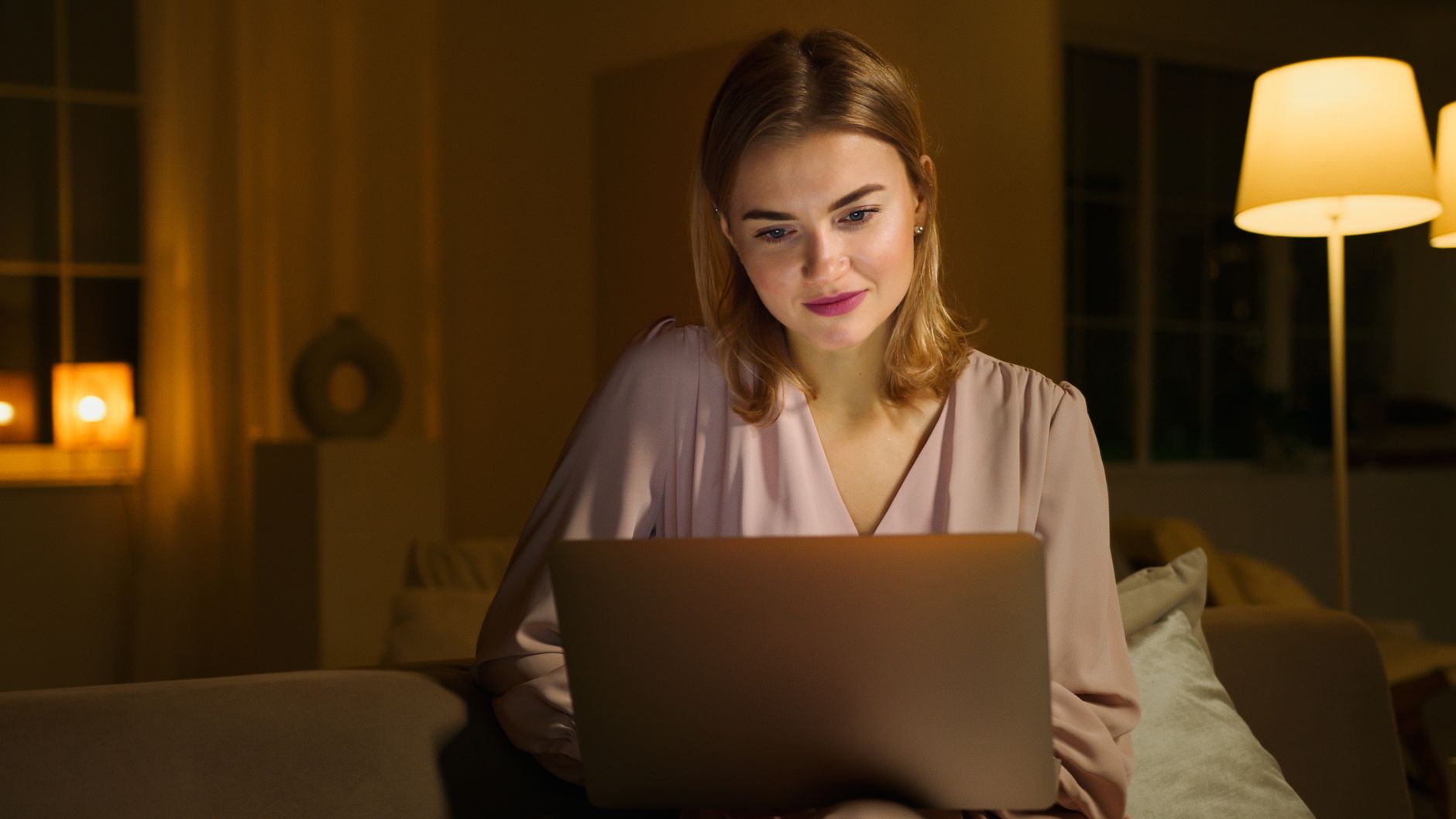 Glimlachende vrouw kijkt op de bank in een warm verlichte woonkamer naar haar laptop