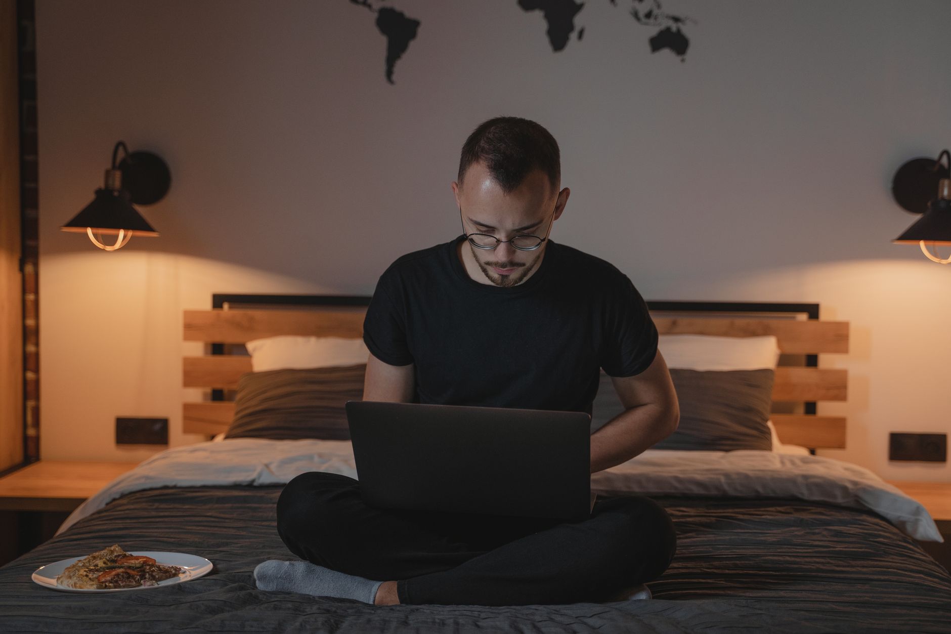 Man met bril zit met gekruiste benen op bed met een laptop op schoot, met een wereldkaart aan de muur